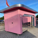 Pink kiosk with a red flag on top, on a paved area with a clear blue sky.