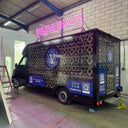 Black van with decorative patterns and a blue logo in a garage with 'Rum Bar' sign.